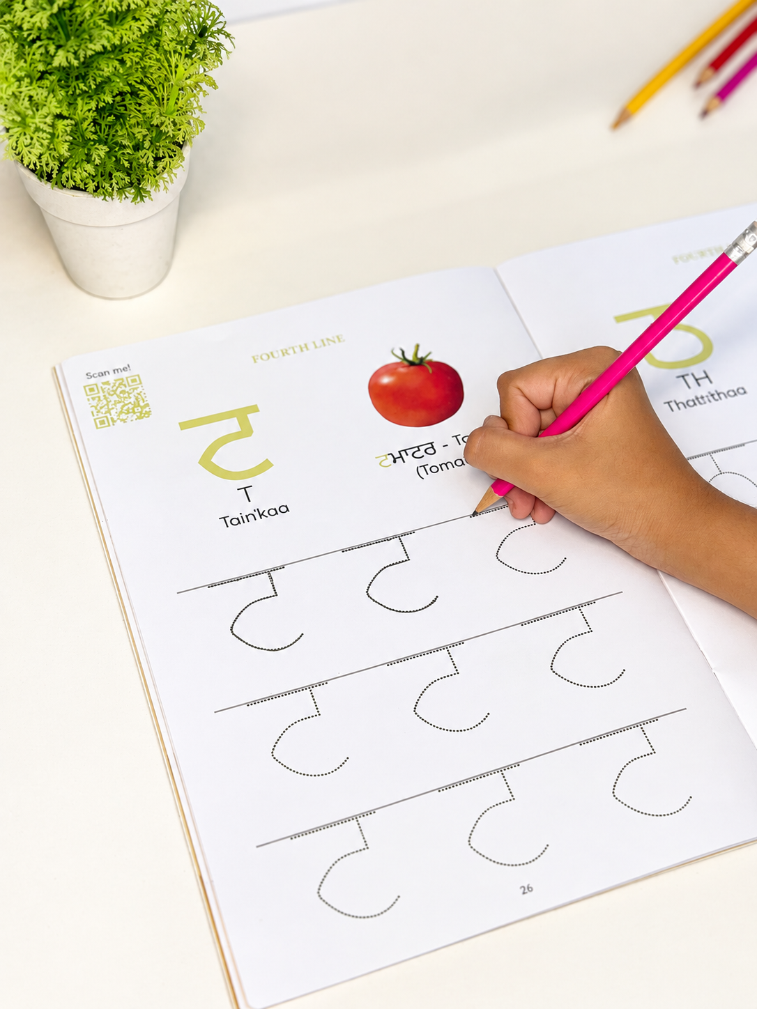 A child's hand holding a pink pencil traces a dotted Punjabi letter practice page from an open workbook on a white desk, with a small potted plant and colored pencils in the background.