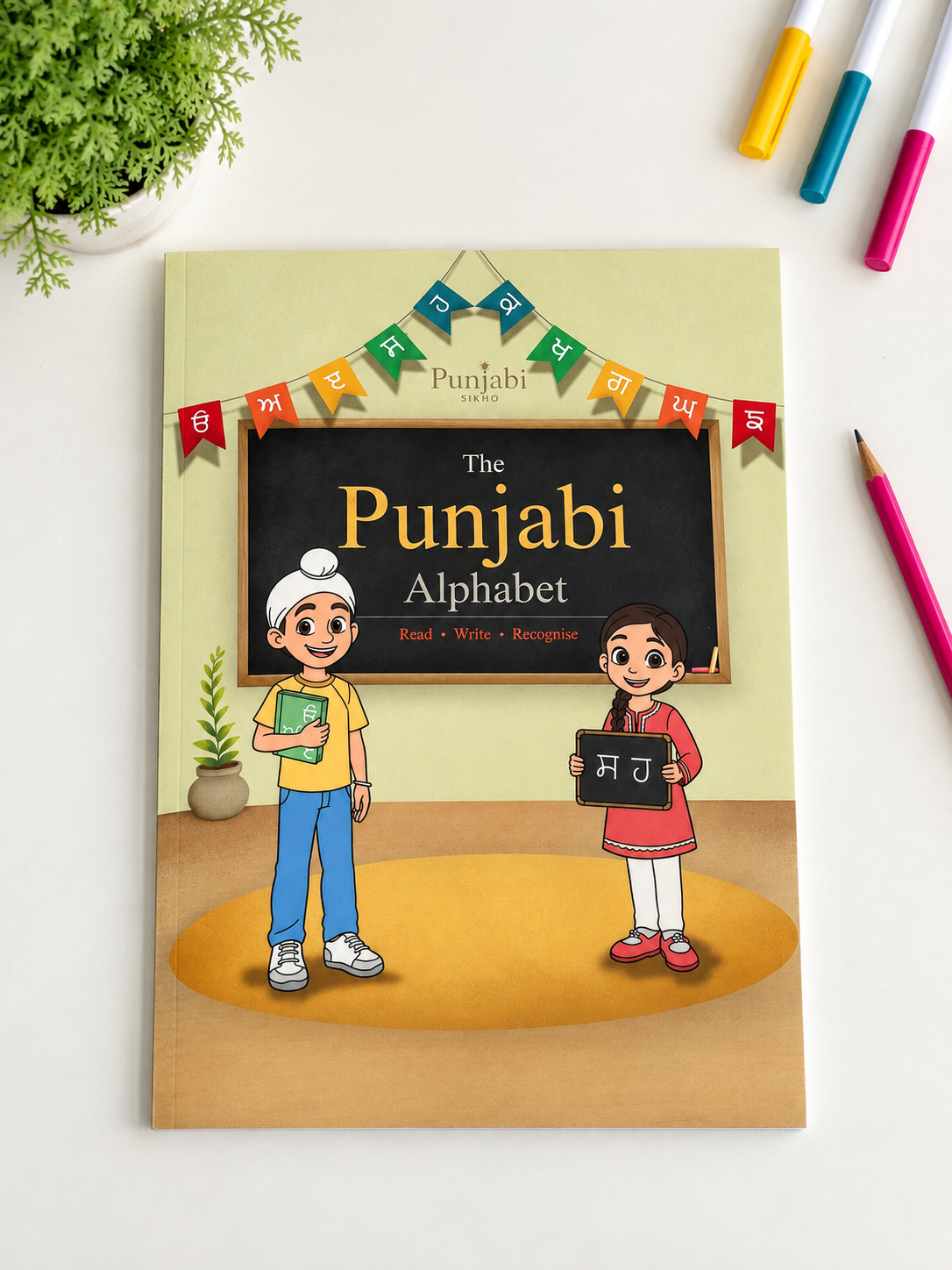 The Punjabi Alphabet book by Punjabi Press, featuring a colorful cartoon illustration of a boy and a girl in front of a blackboard on the cover, displayed on a white desk with colored pencils and a plant.