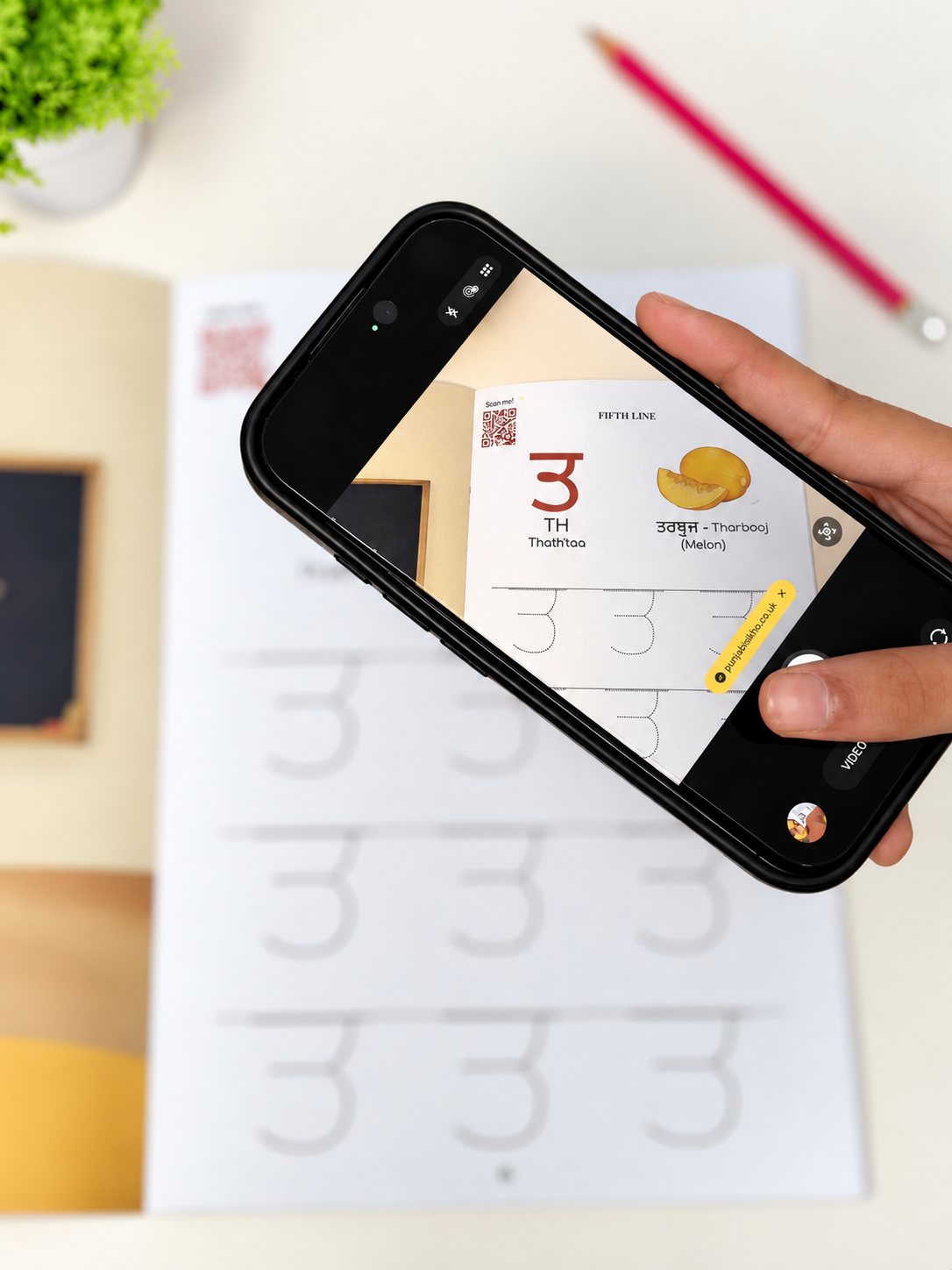 A hand holds a black smartphone over an open practice workbook, using the phone camera to scan or view a page with the letter 'TH' and a small illustration. The scene is a clean desk setup with a pencil and small plant in the background.